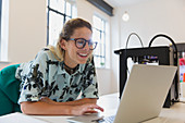 Smiling female designer working at laptop next to 3D printer