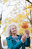Curious senior woman holding orange autumn leaf in park