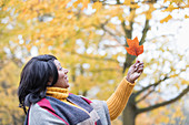 Curious woman holding orange autumn leaf below tree in park