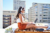 Young woman with headphones using laptop on sunny balcony