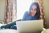 Teenage girl using laptop on bed in sunny bedroom