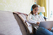 Teenage girl with headphones using laptop on sofa