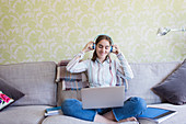 Teenage girl with headphones and laptop on living room sofa