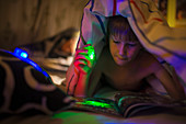 Boy with green flashlight reading magazine under blanket