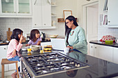 Mother working at laptop, daughters eat breakfast
