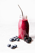 Smoothie in a glass bottle, metal straw, blueberries and blackberries on a marble surface. Front shot.
