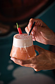 Hands holding glass of foamy drink decorated with straw and chili pepper