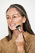 A woman with salt-and-pepper hair with a make-up brush