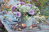 Pastellfarbener Herbststrauß mit Chrysanthemen, Hortensienblüten und Johanniskraut 'Magical Pink Giant', Gräser und Früchte vom wilden Wein liegen