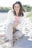 A long-haired woman sitting in the sand on the beach wearing a light jumper and trousers