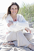 A long-haired woman sitting in the sand on the beach wearing a light jumper and trousers