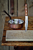 An arrangement of a wooden chopping board, a knife and a saucepan