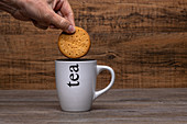Person holding cookie over white mug with hot coffee