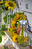A bottle with fennel umbels and marigold flowers with an autumnal wreath behind it