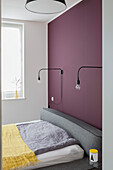 Double bed with grey headboard against plum-coloured wall with reading lamps
