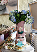 Etagere aus gestapelten Tassen vor Blechkanne mit blauen Hortensien