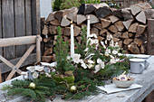 Weihnachts-Tischdeko auf der Terrasse: Kerzen, Zuckerhutfichten in Filztopf, Tannenzweige, Silbertaler und Christbaumkugeln, Brennholzstapel im Hintergrund