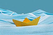 Yellow origami boat on sea of disposable blue face masks