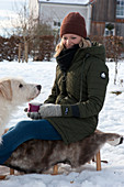 Woman with gloves sitting in a snowy garden on a sled with fur, holding a cup with hot mulled cider wrapped in felt, next to her dog Hera