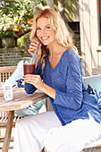 A blonde woman wearing a blue shirt and white trousers with a drink