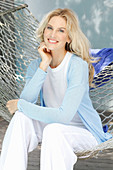 A blonde woman wearing a white top and a light blue cardigan sitting in a hammock