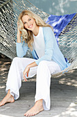 A blonde woman wearing a white top and a light blue cardigan sitting in a hammock