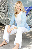 A blonde woman wearing a white top and a light blue cardigan sitting in a hammock