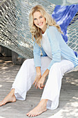 A blonde woman wearing a white top and a light blue cardigan sitting in a hammock