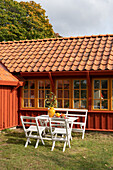 Terrassenplatz mit weißen Möbeln vor rot-braunem Holzhaus mit Sprossenfenstern