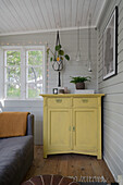 Yellow chest of drawers in the corner of a room, above it macramé flower hanging plants and lanterns