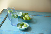 Mojito cocktails in blue drinking glasses with open bottle and bowls of limes, mint and sugar