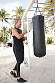 A grey-haired man wearing black sports clothing using a punching bag