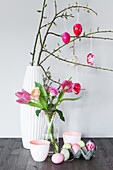 Branches with marbled Easter eggs in a white vase, in front of it a glass vase with tulips