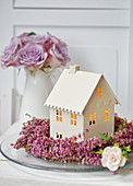 House-shaped tealight lantern in wreath of heather and vase of purple roses