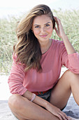 A young, long-haired woman wearing a pink blouse and shorts sitting in the sand