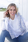 A long-haired blonde woman wearing a white shirt blouse and jeans with a jumper over her shoulders