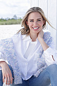 A long-haired blonde woman wearing a white shirt blouse and jeans with a jumper over her shoulders