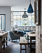 Open plan kitchen diner with mismatching beaded chandeliers, rustic bench and mid century armchair, floors white-washed oak