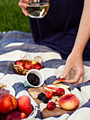 A woman holding a glass of white wine, reaching for a slice of peach, picnic with fruit and cheese