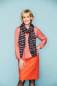 A blonde woman with short hair wearing a pink blouse with a scarf and an orange skirt