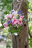 Kleiner Strauß aus Rosen, Akelei, Ehrenpreis und Blüten vom Wiesenkümmel in Glas mit Graskranz an Pfosten gehängt