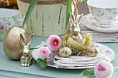 Osterteller mit goldenem Osterhasen in Eierschale auf Moos, Blüten von Tausendschön, Ostereier naturfarben und golden