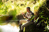 Man fly fishing and using smartphone at sunny riverbank