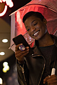 Happy young woman using smartphone under umbrella at night