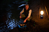 Happy man cooking on camping stove at riverbank at night