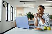 Thoughtful young businesswoman drinking coffee at laptop