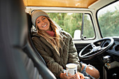 Happy young woman drinking coffee at steering wheel in van