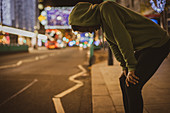 Female runner resting with hands on knees on street at night