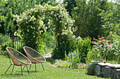 Rose arch with rambler rose 'Ghislaine de Feligonde', Acapulco chair on the lawn, cushions on natural stones making a bench, decorative perennial support in the bed