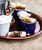 Enamel mugs of hot chocolate, whipped cream, and skewers of roasted marshmallows